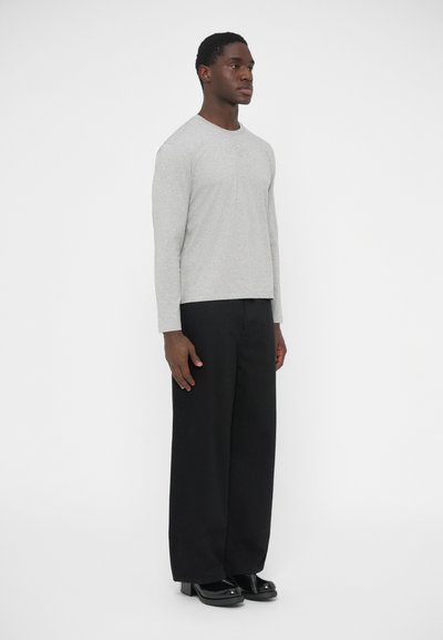 Sivá košeľa s dlhými rukávmi má okrúhly výstrih a jemný predný šev. Je kombinovaná s voľnými čiernymi nohavicami a lesklými čiernymi topánkami.