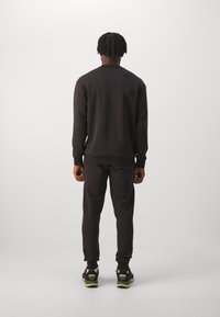 Felpa nera e pantaloni della tuta neri, entrambi con polsini a costine e una morbida texture. Le scarpe sportive nere e verdi completano l'outfit.