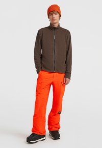 Bruine zip-up jas over feloranje sneeuwbroeken met cargozakken, gecombineerd met een bijpassende oranje beanie en zwarte sneeuwschoenen.