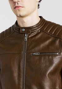Giacca da moto in pelle marrone con colletto strutturato, zip frontale verticale e tasca sul petto con zip. Presenta dettagli testurizzati sulle spalle.