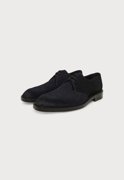 Paire de chaussures habillées pour hommes noires texturées à lacets, avec petits talons et semelle lisse sur un fond uni.