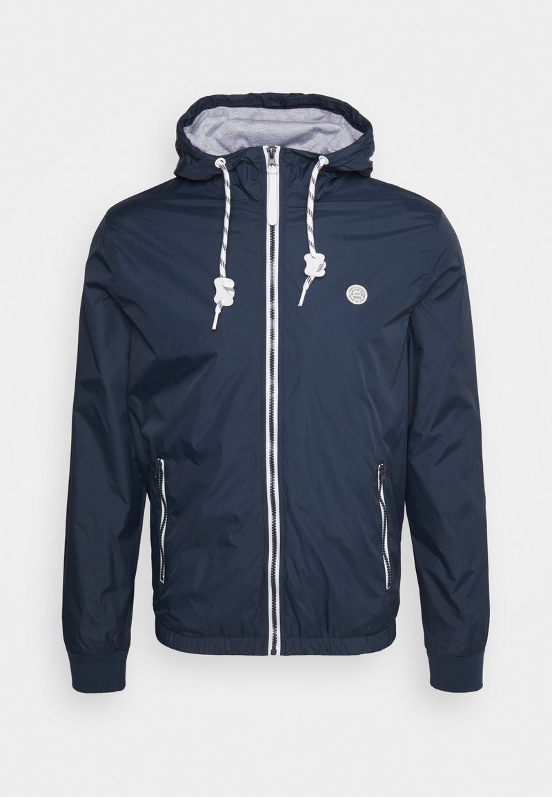 Veste à capuche bleu marine avec fermeture éclair blanche et cordons, poignets élastiques, poches zippées et petit logo rond sur la poitrine.