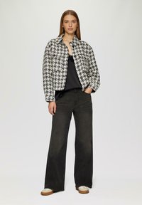 Veste à motif pied-de-poule noir et blanc avec des boutons, associée à un haut noir et un jean large foncé. Les chaussures présentent des accents crème et tan.