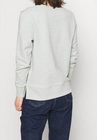 Jasnoszary sweter z okrągłym dekoltem, ściągaczami przy rękawach i u dołu, z bocznymi rozporkami i miękką teksturą. Noszony z ciemnoniebieskimi dżinsami.