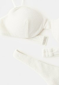 Haut et bas de bikini en tissu texturé blanc avec fermoirs ajustables, posés à plat sur un fond blanc.