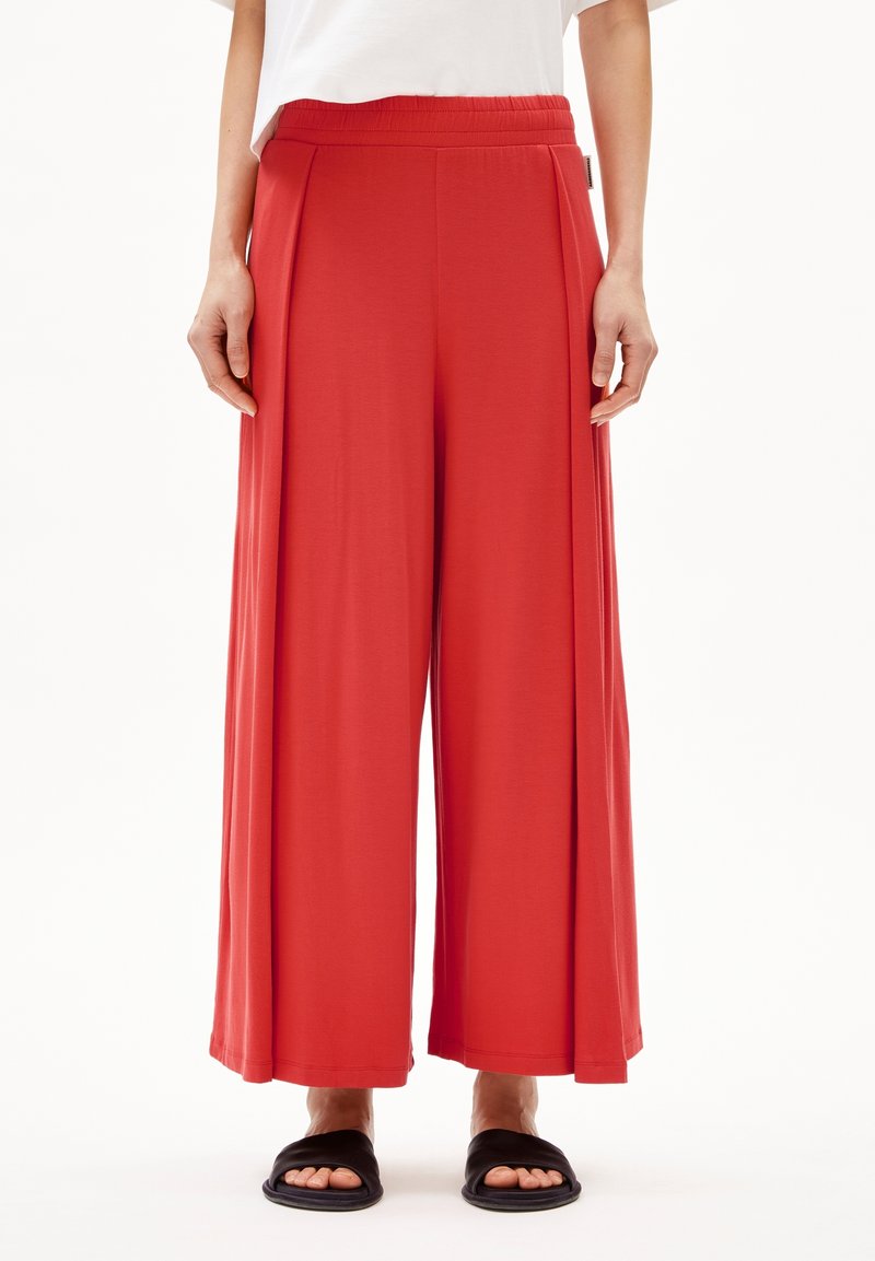 Personne portant un pantalon rouge ample à jambes larges, un haut blanc et des sandales noires ouvertes aux orteils, debout devant un fond blanc.