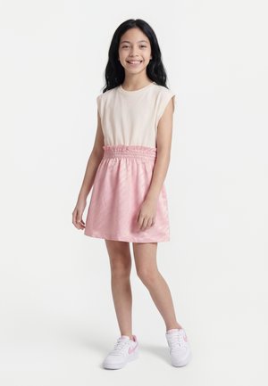 Meisje staand en glimlachend, gekleed in een mouwloos crèmekleurig topje, een roze rok met geplooide taille, en witte sneakers met roze accenten.