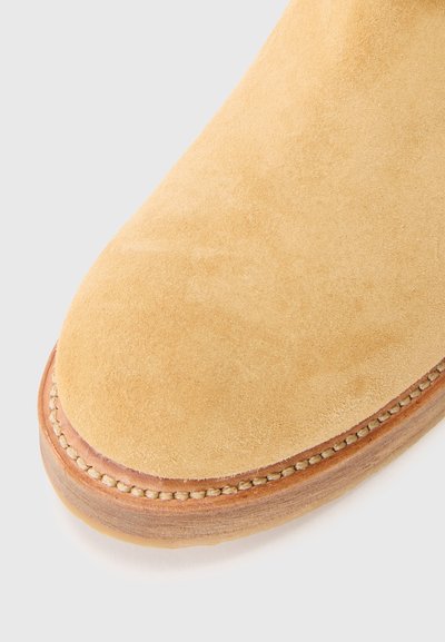 Bota de ante en amarillo claro con puntera redondeada. Presenta una suela de madera cosida con un acento beige alrededor del borde. Textura suave.