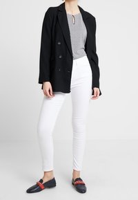 Schwarzer zweireihiger Blazer über einem gemusterten Top, kombiniert mit weißen Skinny-Jeans und marineblauen Mokassins mit roten Akzenten.