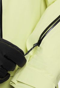 Wasserdichte gelbe Jacke mit schwarzem Reißverschluss und Kordelzug. Das Material hat eine glatte Textur. Eine behandschuhte Hand zieht den elastischen Kordelzug an.