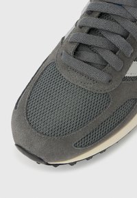 Gros plan sur l'avant d'une chaussure de sport grise avec un tissu en mesh et en daim, des lacets épais et une bande latérale blanche sur fond blanc.