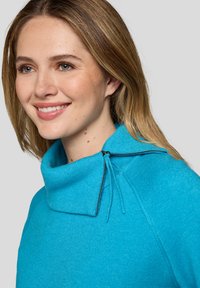 Lächelnde Frau mit hellbraunen Haaren, die einen leuchtend blauen Pullover mit hohem Kragen trägt, der einen seitlichen Reißverschluss und Kordelzüge hat.