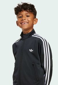Smiling boy with curly hair wearing a black Adidas zip-up jacket with white stripes on sleeves and logo on chest against a plain background.
