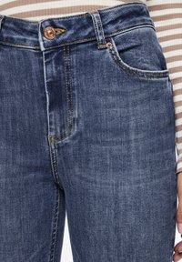 Høyt liv blå denimjeans med kobberknapp, kontrasterende stikking og en slitt tekstur. Har en frontlomme og tapered passform.