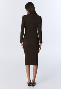 Langärmliges, geripptes braunes Kleid, figurbetonte Silhouette, knielang, mit rundem Ausschnitt und Rücken-Schlitz. Schwarze, mit Nieten besetzte flache Schuhe ergänzen den Look.