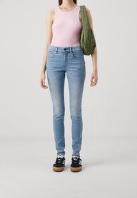 Jeans ajustados de color azul claro con un acabado desgastado, combinados con una camiseta de tirantes de canalé rosa y zapatillas negras con rayas blancas. Bolsa acolchada verde.