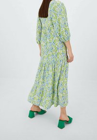 Frau mit glattem braunem Haar, die ein langes floral grünen-blaues Kleid und grüne Blockabsatzsandalen trägt, stehend vor einem schlichten weißen Hintergrund.