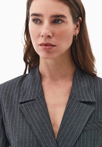 Blazer gris de rayas con solapas de muesca, un bolsillo en el pecho y un cuello en V profundo. Tela suave y ajustada con un corte elegante y moderno.