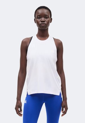 Vrouw draagt een wit mouwloos sporttopje en blauwe legging, staand tegen een effen lichte achtergrond.