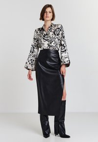 Donna che indossa una blusa floreale in bianco e nero, una gonna di pelle nera alta in vita con spacco laterale e stivali neri con tacco, in piedi contro uno sfondo bianco.