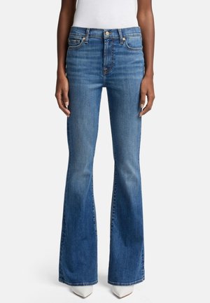 Jean slim fit en denim bleu moyen avec taille haute et jambes évasées, porté avec des chaussures blanches à bout pointu et un haut blanc.