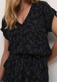 Top negro de cuello en V con mangas cortas, que presenta un patrón floral en tonos de azul y verde, y un detalle de cintura atada para darle forma.