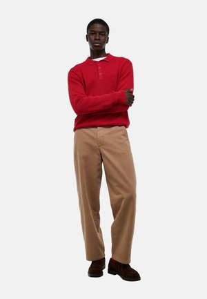 Jeune homme debout, les bras croisés, portant un pull polo rouge à manches longues, un pantalon beige et des chaussures marron sur un fond blanc uni.