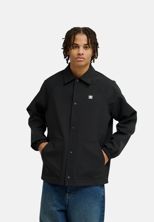 Junge Person mit geflochtenem Haar, die eine schwarze, geknöpfte Jacke mit Fronttaschen und blaue Jeans trägt, steht mit den Händen in den Taschen.