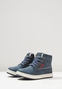 Botas impermeables azules hechas de cuero texturizado, con una suela de goma blanca, cordones a rayas azules y blancas, y un forro de tela suave.