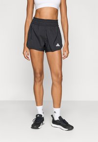 Egy nő fekete Adidas sportnadrágot, fehér sportruhát, fehér Adidas zoknit és fekete tornacipőt visel, és egy egyszerű háttér előtt áll.
