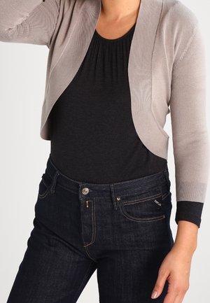 Femme portant un haut noir, un cardigan court beige clair et un jean bleu foncé, la main posée sur la hanche.