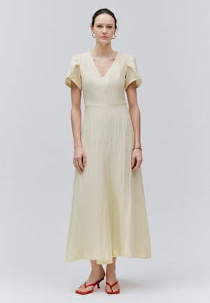 Mujer de pie vestida con un vestido midi color crema con cuello en V, mangas cortas abullonadas y sandalias de tacón rojas, sobre un fondo claro y liso.