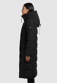Schwarze, gepolsterte Jacke mit hohem Kragen, Kapuze und Reißverschlusstaschen; verfügt über eine gesteppten Textur und ein Logo-Patch am Ärmel.