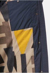 Chaqueta de forro polar con una mezcla de patrones geométricos en beige, gris y amarillo, con un panel de tela azul marino y detalles de botones a presión.