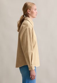 Beżowa koszula sztruksowa z długimi rękawami, zaostrzonym kołnierzykiem i mankietami na guziki, w połączeniu z jasnoniebieskimi dżinsami. Miękki, fakturowany materiał.