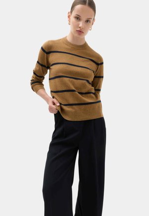 Jonge vrouw met haar naar achteren gebonden, gekleed in een bruine trui met zwarte horizontale strepen en zwarte broek met wijde pijpen.