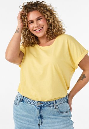 Smilende kvinde med krøllet hår iført gul t-shirt og lyseblåt denimskørt, den ene hånd rører ved håret, tatoveringer synlige på underarmene.