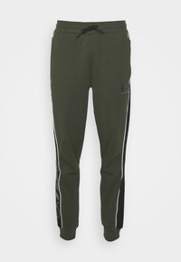 Olivově zelené tepláky s černými bočními pruhy a bílým lemováním; mají stahovací pas a logo v přední části.