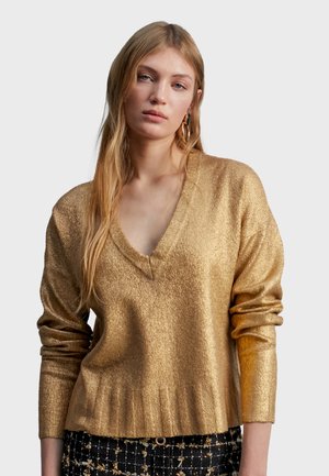 Pull en V doré, avec une finition texturée, à manches longues et ourlet côtelé. Porté de manière décontractée, associé à un pantalon à motifs.