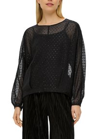 Schwarze, durchsichtige Bluse mit Rundhalsausschnitt und langen Puffärmeln, die mit strukturierten, perlenartigen Mustern versehen ist. Getragen mit schwarzen plissierten Hosen.