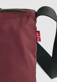 Bordeaux stofpose med lynlås, med en sort rem og en rød label. Struktureret overflade, kompakt design og minimalistiske detaljer.