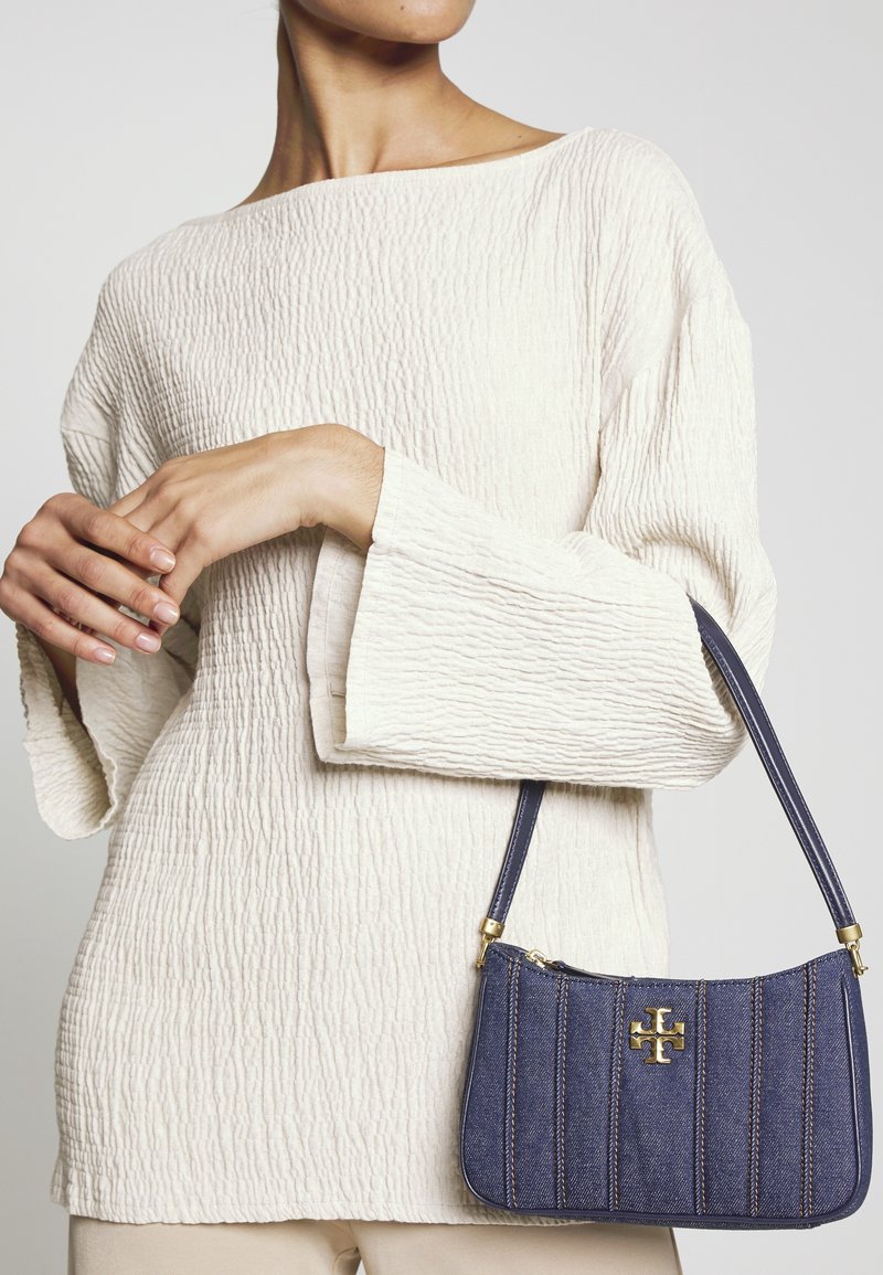 Femme portant un haut texturé à manches longues blanc cassé tenant un sac à main bleu marine avec un logo doré et des détails de couture sur la sangle.
