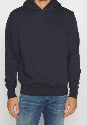 Homme portant un sweat à capuche bleu marine avec un petit logo sur la poitrine et un jean bleu, debout devant un fond clair uni.