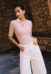 Tumejuustega naine, kes kannab triibulist lühikese varrukaga särki ja valgeid laia säärega pükse, seisab käed taskus kiviseina taustal.