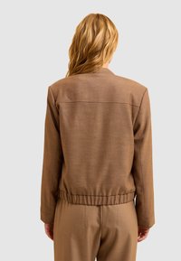 Femme aux cheveux blonds ondulés portant une veste marron et un pantalon assorti, de dos contre un fond gris clair uni.