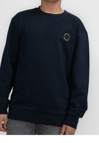 Sweat-shirt bleu marine en coton avec des manches longues, des poignets côtelés et une petite pièce noire sur la poitrine arborant un motif circulaire.
