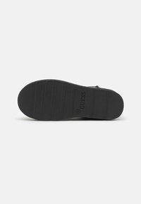 Suela de zapato de goma negra con patrón estriado, logotipo "GUESS" en relieve y marcado de talla. Diseño plano con una textura suave.