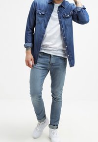 Camicia di denim blu scuro, t-shirt bianca sotto, abbinata a jeans skinny blu chiaro e sneakers bianche, con due tasche sul petto.