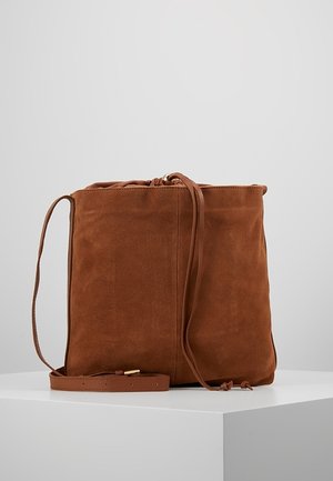 Sac bourse en daim marron avec bandoulière réglable et fermeture à cordon, présenté sur une surface blanche contre un fond gris.