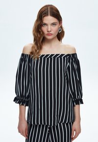 Sort og hvid stribet top med off-shoulder udskæring og klokkeærmer. Har volanger i manchetten og en løs pasform, lavet af letvæv.
